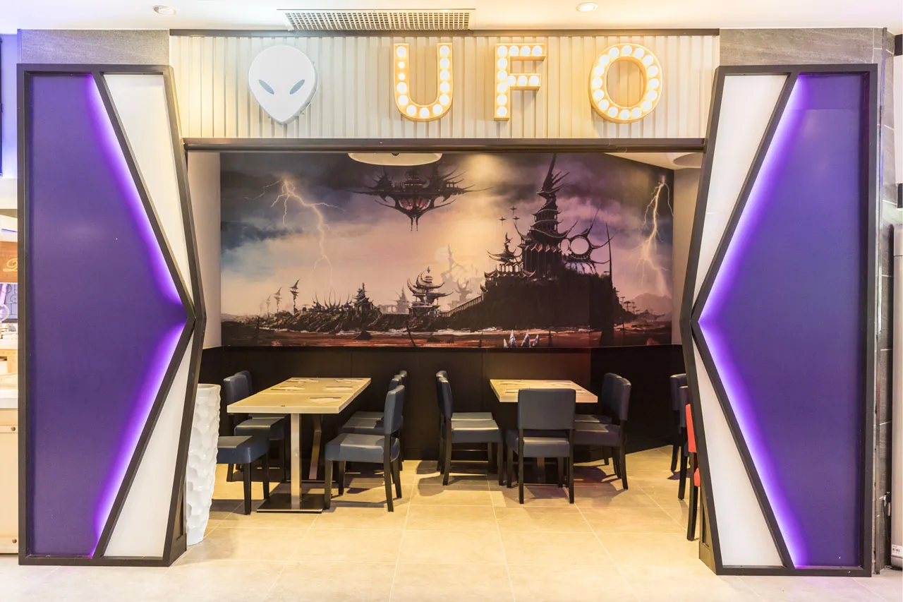 Alcôve de tables sous une enseigne lumineuse « UFO » et une fresque de cité futuriste